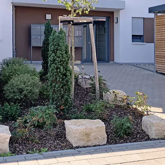 Vorgarten mit Beetanlage, kleinen Bäumen und dekorativen Natursteinen vor einem modernen Gebäude.