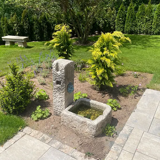 Garten mit Natursteinbrunnen in einem bepflanzten Beet, umgeben von Rasenflächen und einer Sitzbank im Hintergrund.