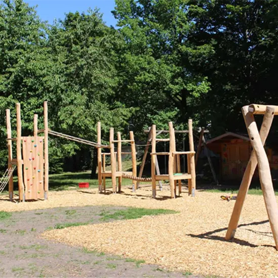 Spielplatz aus natürlichen Holzelementen mit Kletter- und Balanciermöglichkeiten, umgeben von Bäumen und Holzschnitzeln als Bodenbelag.