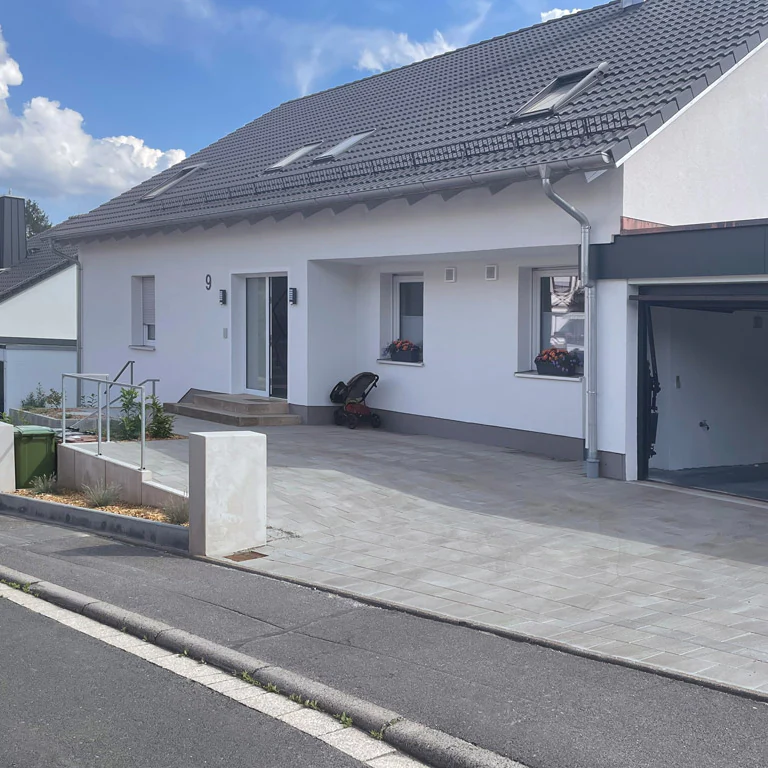 Modern gepflasterte Einfahrt mit hellen Steinen vor einem Wohnhaus, eingefasst von Betonpfeilern und einer kleinen Treppe mit Geländer.